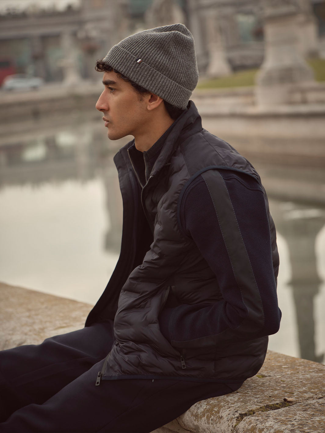 Mann in dunkler Kleidung und Mütze sitzt auf einer Mauer und hat die Hände in die Jackentaschen gesteckt