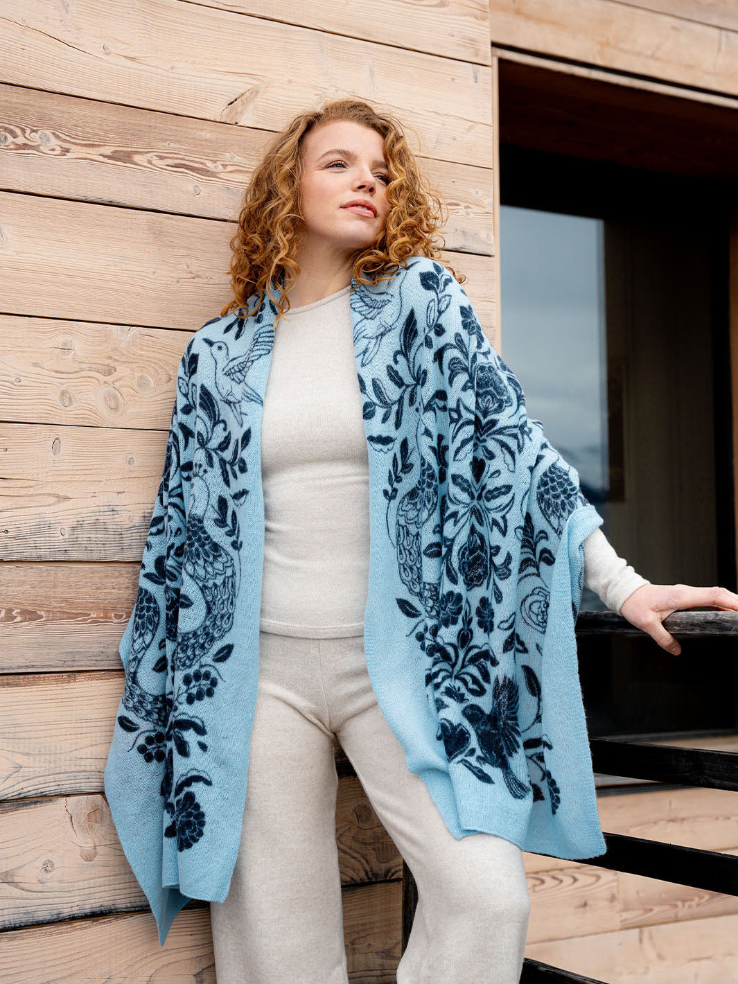 Frau mit warmer Stola 684123 um die Schultern steht an eine Hauswand aus Holz gelehnt da