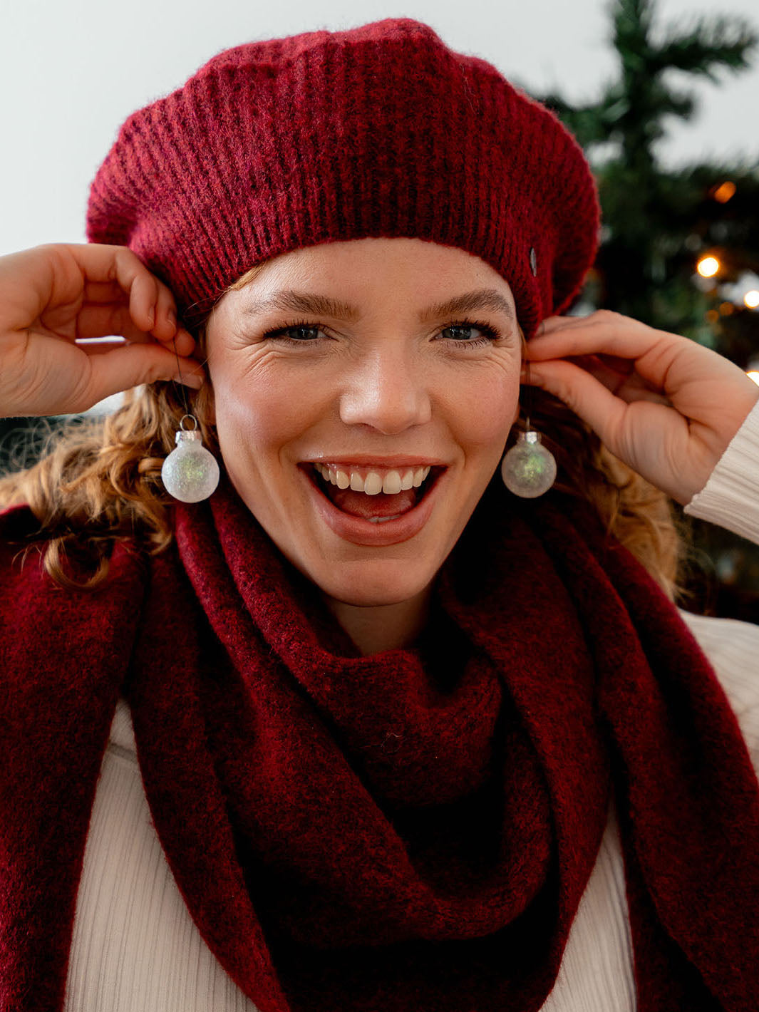 lachende Frau mit Schal und Mütze hält sich kleine Christbaumkugeln als Ohrringe ans Ohr