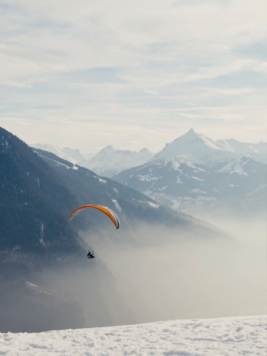 Eine Person fliegt mit einem Paragleitschirm an zwischen verschneiten Bergen.