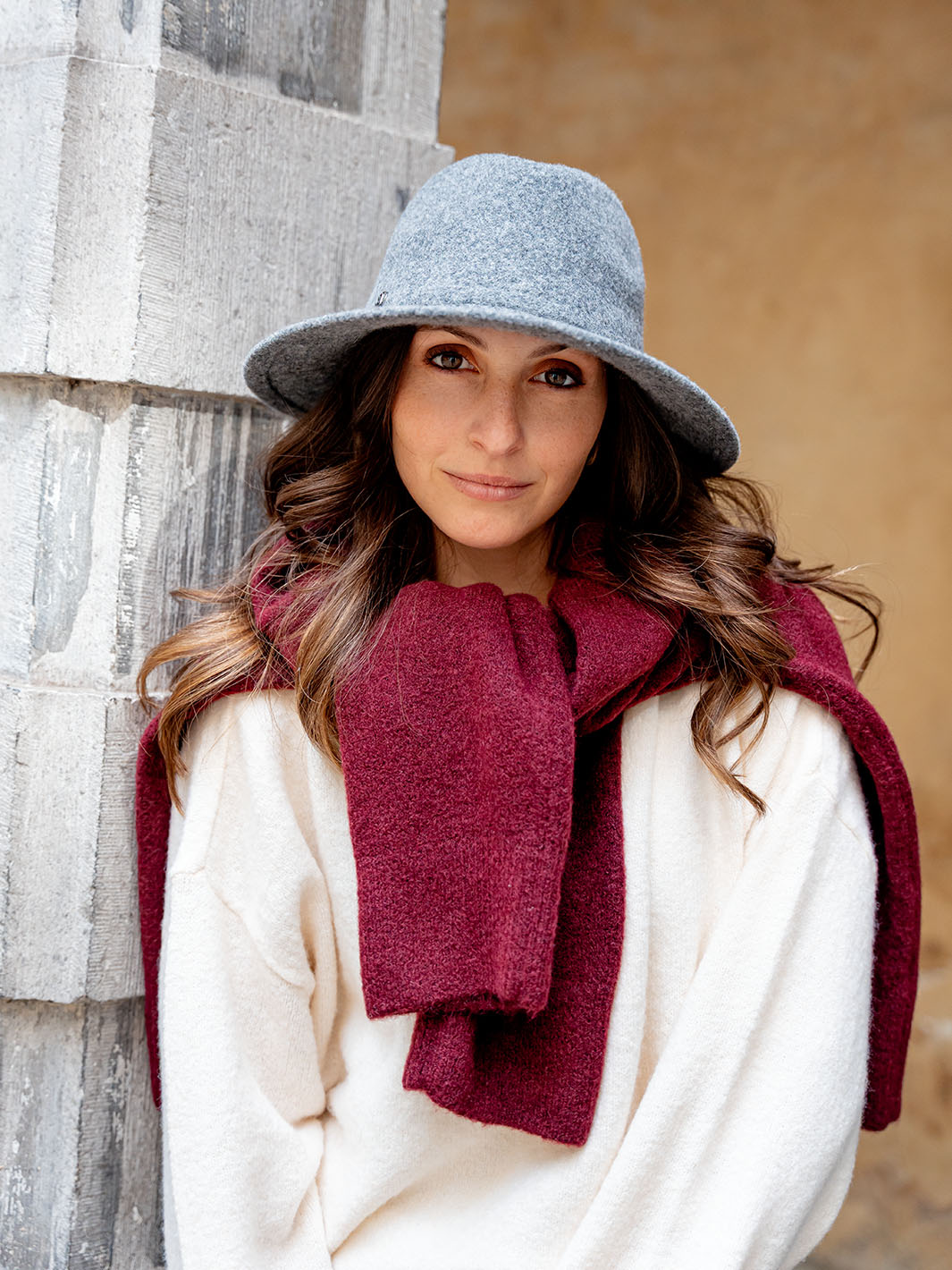 Frau mit LOEVENICH Fedora 672500 steht seitlich an einer Mauer und sieht in die Kamera