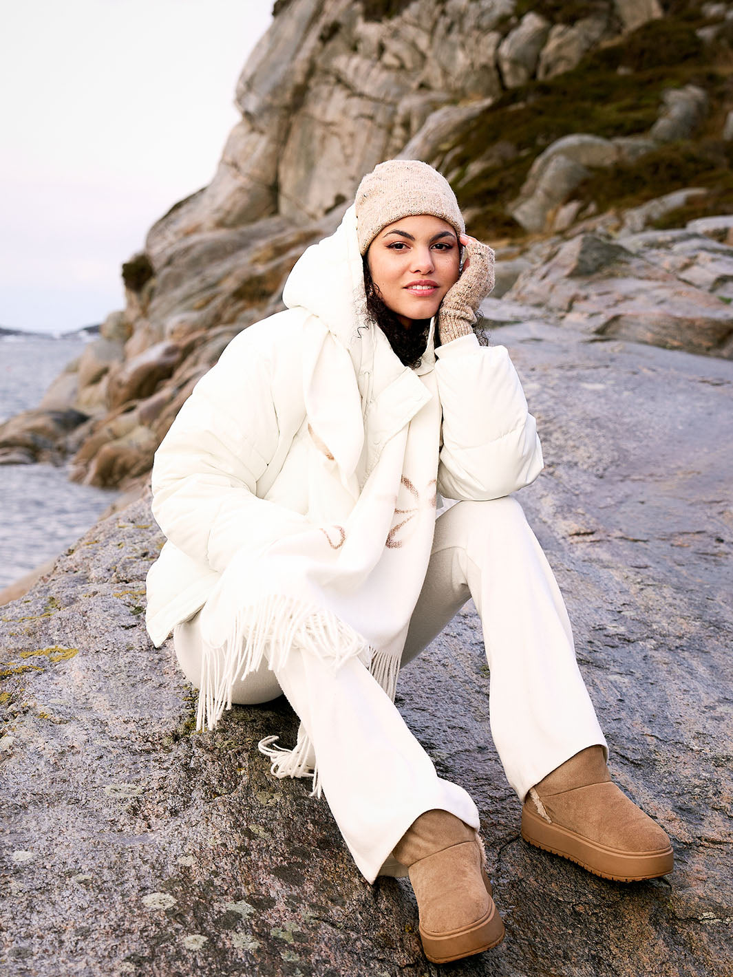 Frau mit Winterjacke, Strickmütze und dazu passenden Stulpen vor Felsen am Meer 