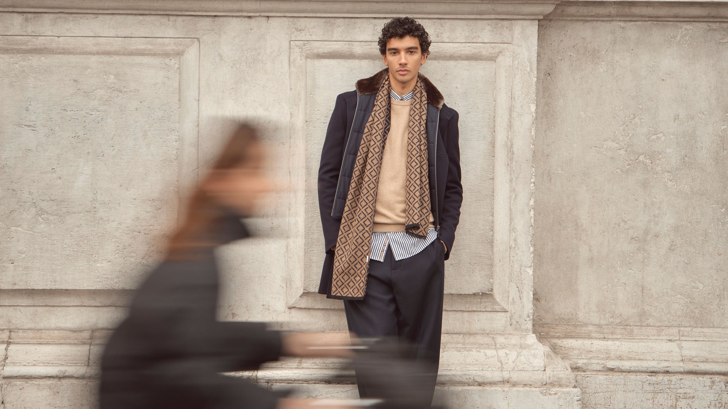 Mann mit Jacke und gemustertem Schal hat die Hände in den Hosentaschen und sieht in die Kamera