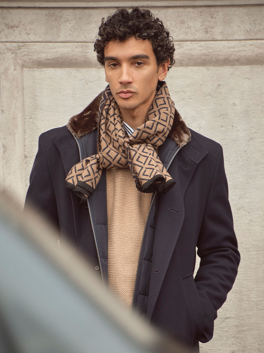 Mann mit Jacke und gemustertem Schal hat die Hände in den Hosentaschen und sieht in die Kamera