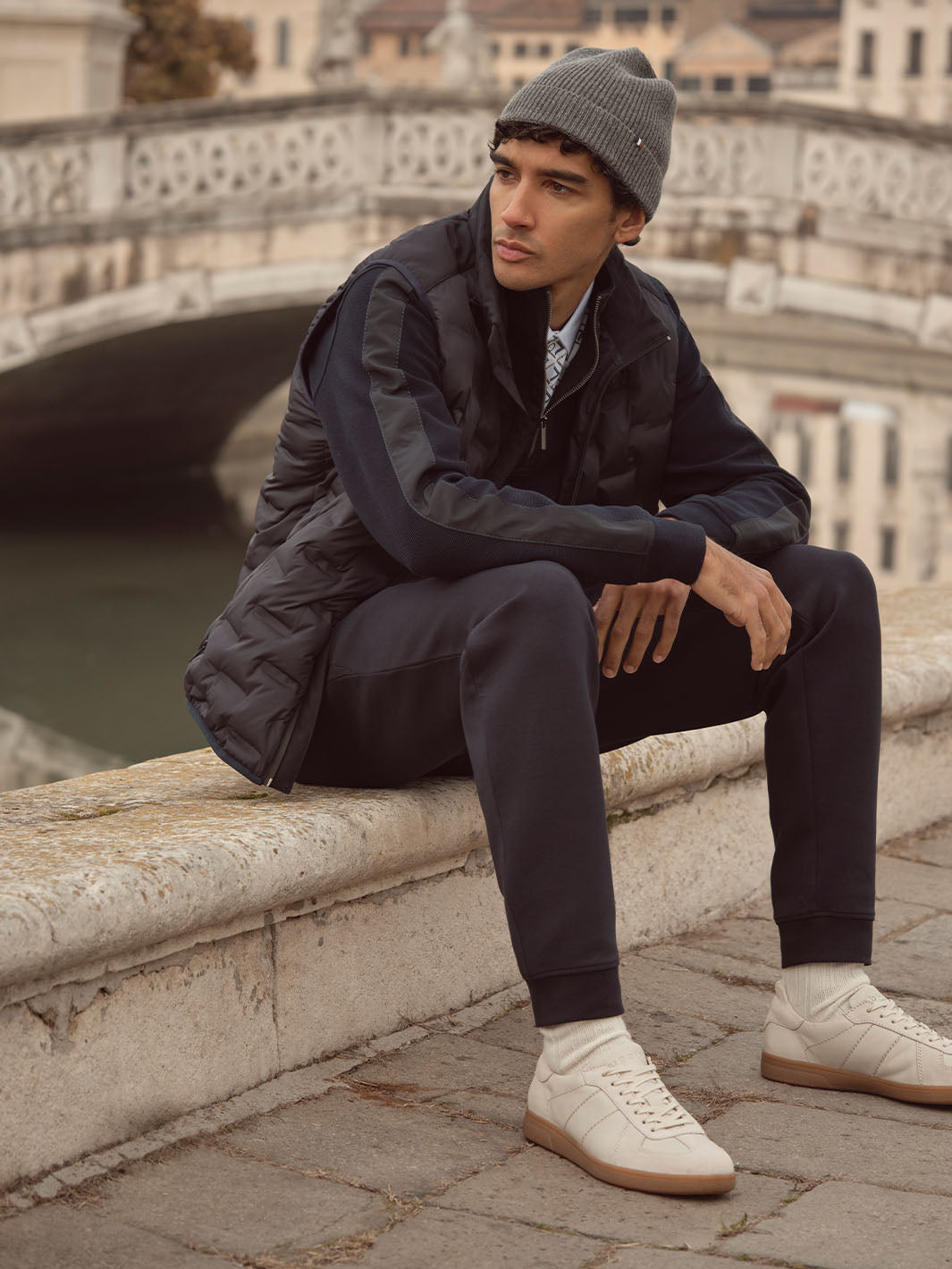 Ein Mann mit bugatti Kleidung sitzt auf einer Brüstung über einem Fluss in der Stadt