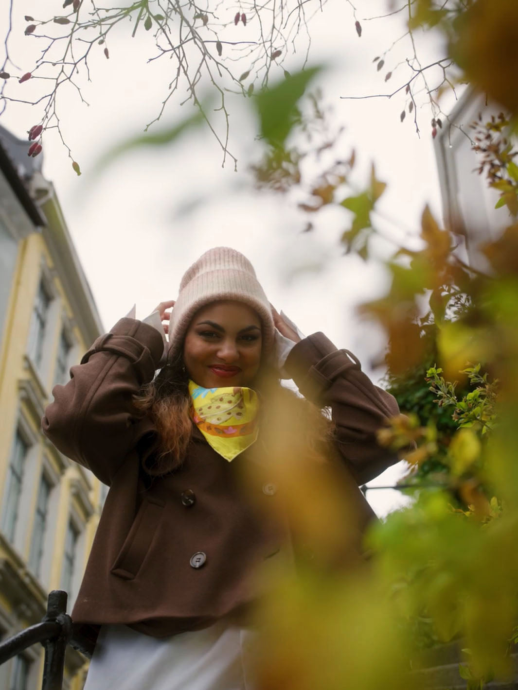 Frau mit gestrickter Wintermütze und bunt gemustertem Seidentuch am Hals durch die Äste eines Busches fotografiert