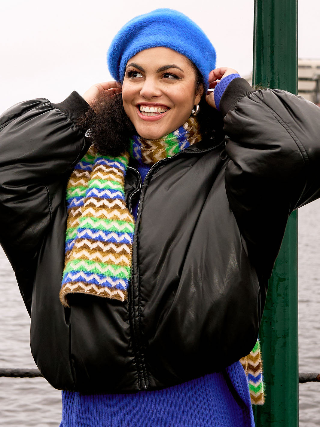 Lachende Frau mit Baskenmütze und dazu passendem Schal steht vor einem Hafen, im Hintergrund Wasser