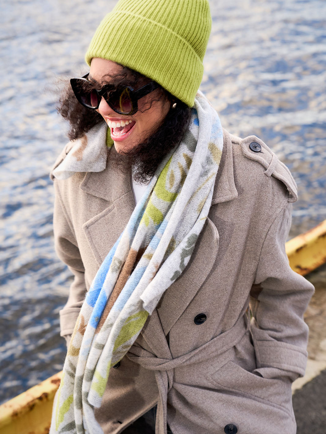 Frau mit Sonnenbrille und grüner Strickmütze am Kopf steht auf einem Steg überm Wasser