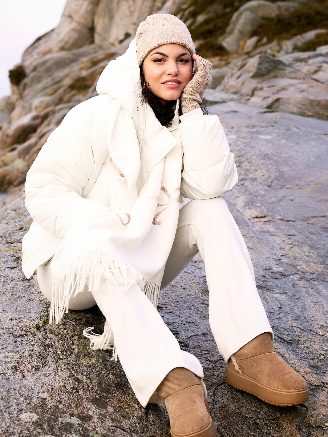 Frau mit Winterjacke, Strickmütze und dazu passenden Stulpen vor Felsen am Meer 