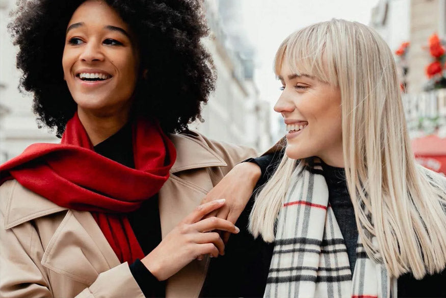 zwei Freundinnen gehen lachend durch die Stadt, eine hat die Arme auf die Schultern der anderen gelegt