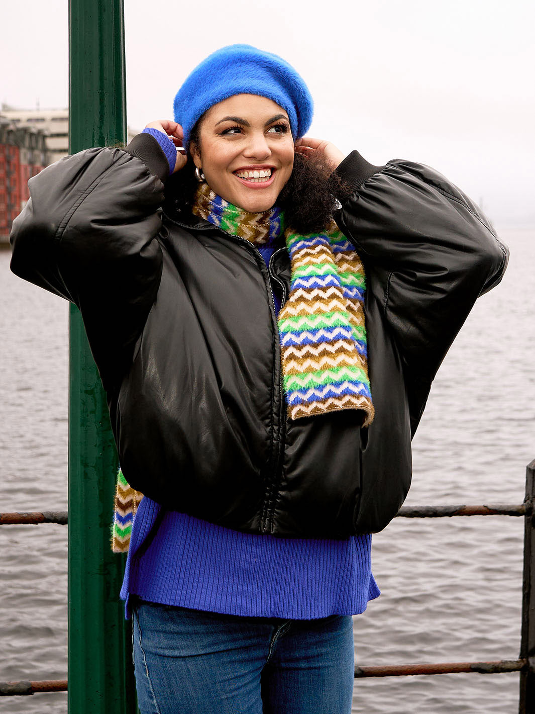 Lachende Frau in warmer Kleidung setzt ihre Baskenmütze auf, im Hintergrund Wasser und Häuser