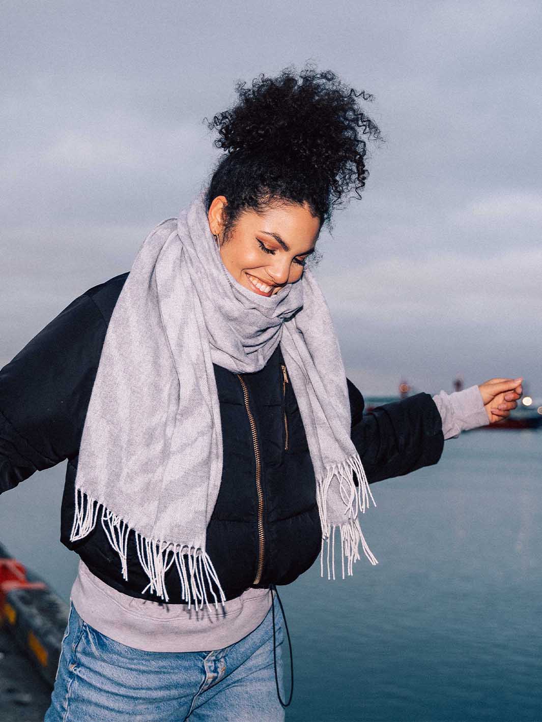 Frau hat sich ihren Schal um den Hals gewickelt und balanciert auf einer Brücke, im Hintergrund das Meer