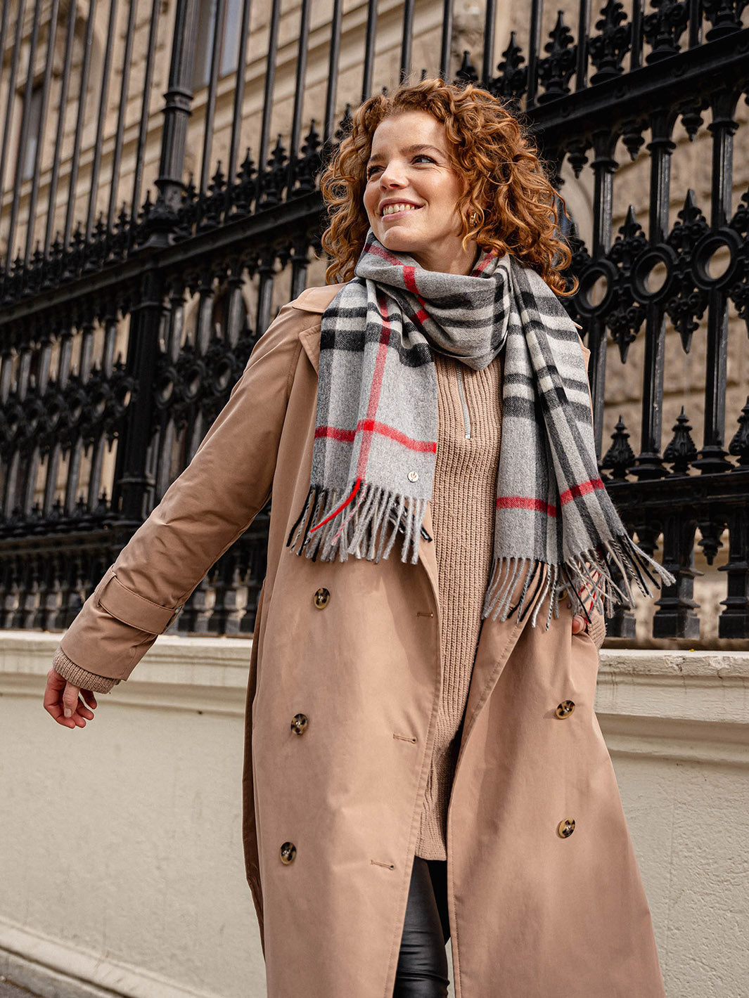 Frau mit Wintermantel und FRAAS Plaid Schal läuft an einem schwarzen Zaun aus Eisen vorbei