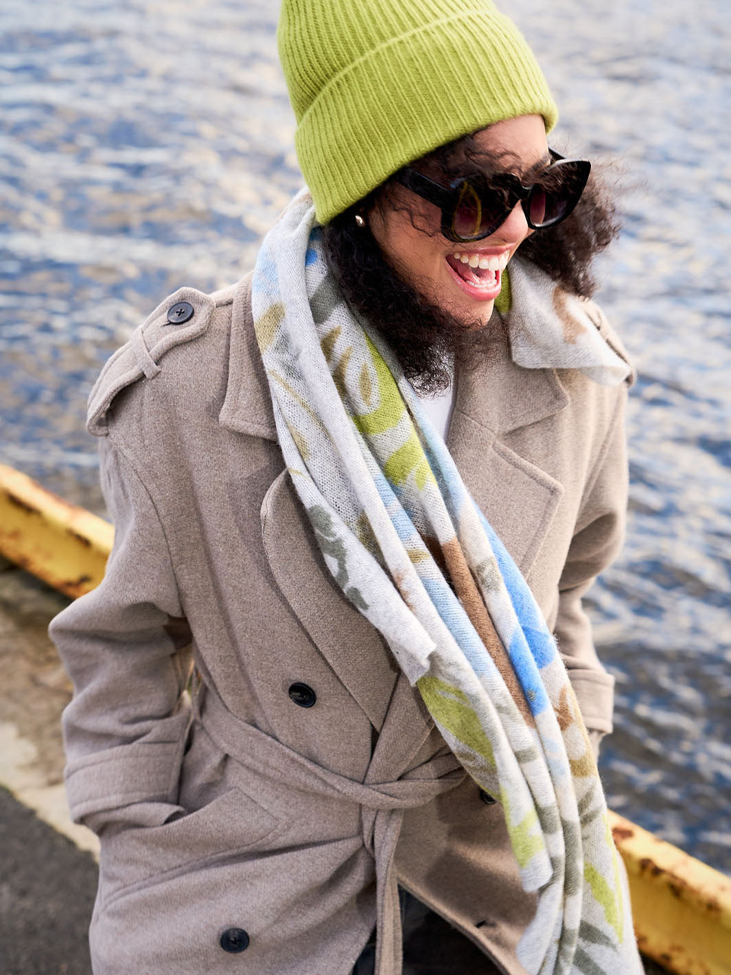 Frau mit Mantel, Schal und Strickmütze steht lachend im Wind, im Hintergrund Wasser