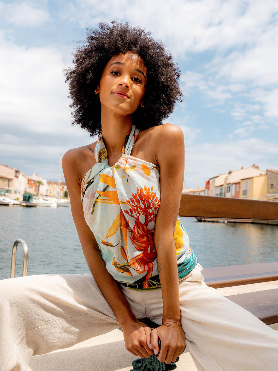 Frau trägt die FRAAS Seidenstola 609063 als Top. sie sitzt auf einer Mauer, hinter ihr das Meer, ein Hafen mit Booten und Häuser.