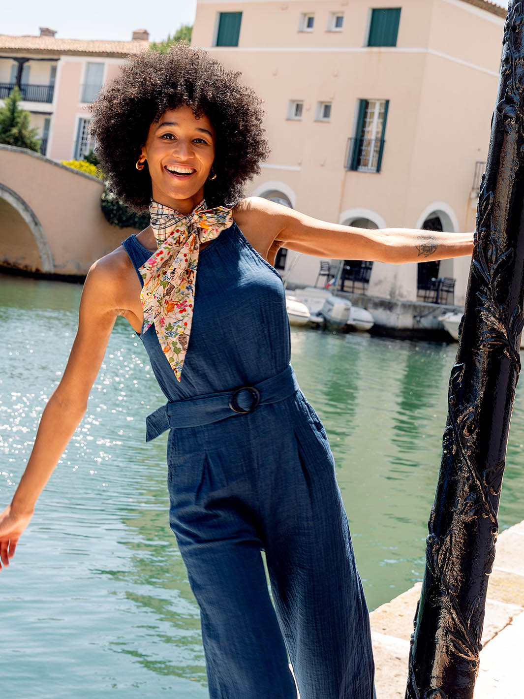 Frau trägt Twilly Seidenschal 612021 mit FRAAS Plaid Karo und Tier-Print als Schleife um den Hals und hängt sich mit dem Arm lachend an eine Laterne. In Hintergrund ein Kanal, Boote und Häuser.