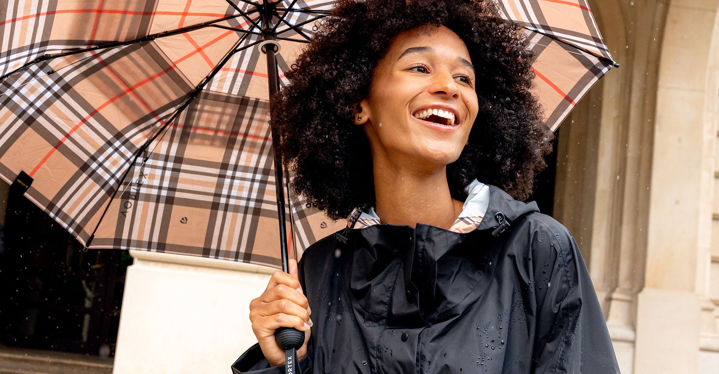 Frau im FRAAS Plaid Regenponcho hält bei Regen lachend einen Regenschirm über sich.