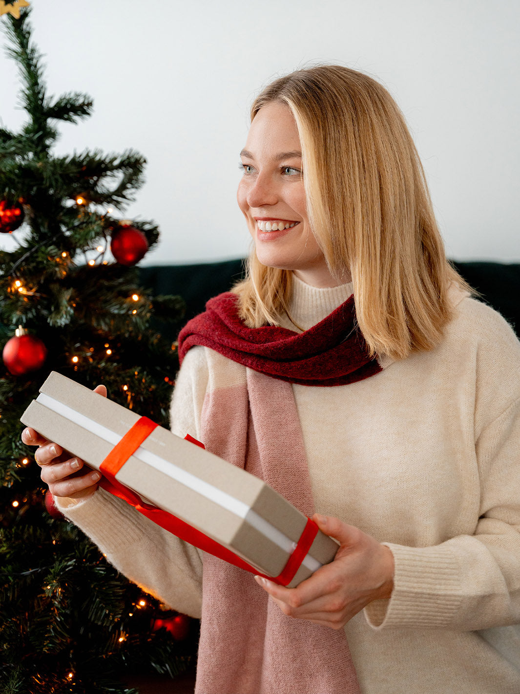 Frau steht vor dem Weihnachtsbaumund hat ein verpacktes Geschenk in der Hand.