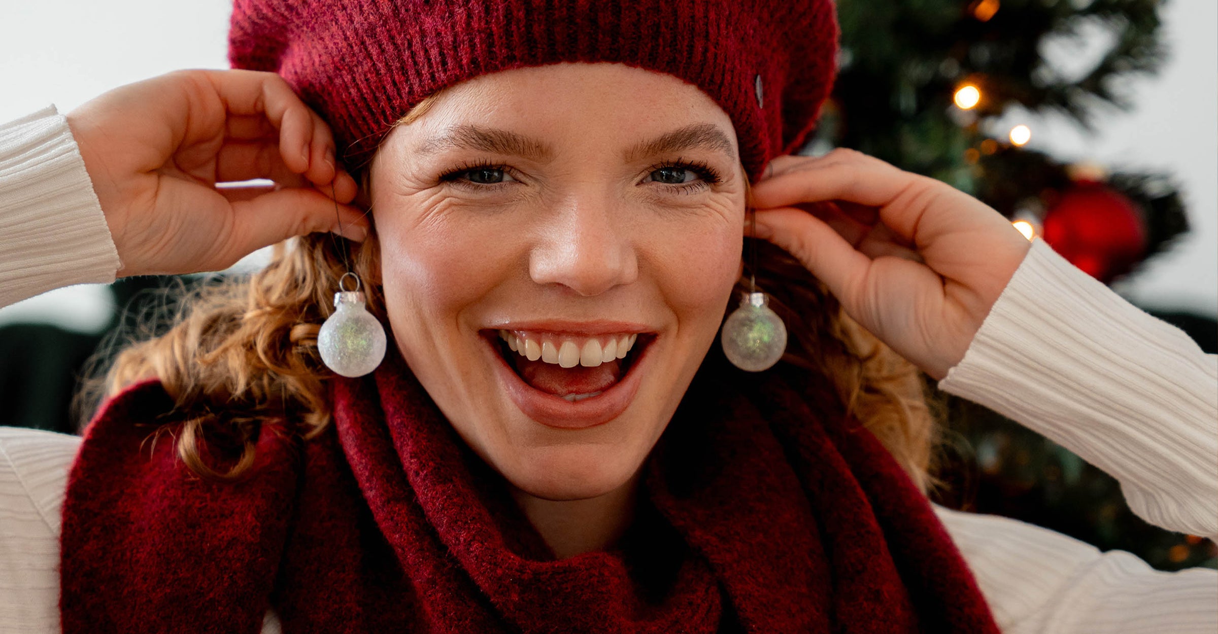Frau mit Schal und Mütze hält sich Christbaumkugeln als Ohrringe an die Ohren, im Hintergrund ein Weihnachtsbaum
