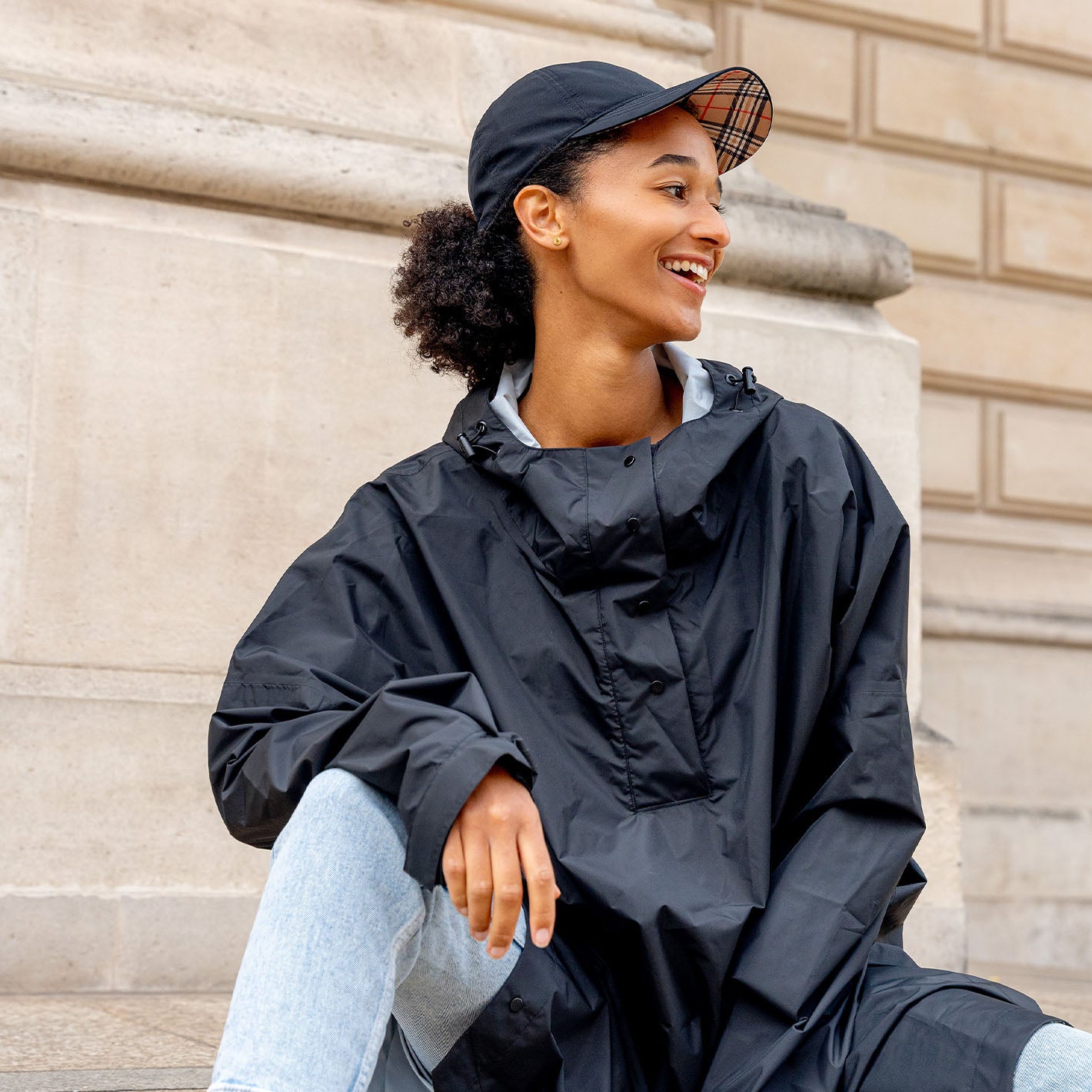 Junge Frau sitzt auf einer Treppe. Sie trägt die FRAAS Plaid Regen-Kappe 602064 und den Regen-Poncho 602801