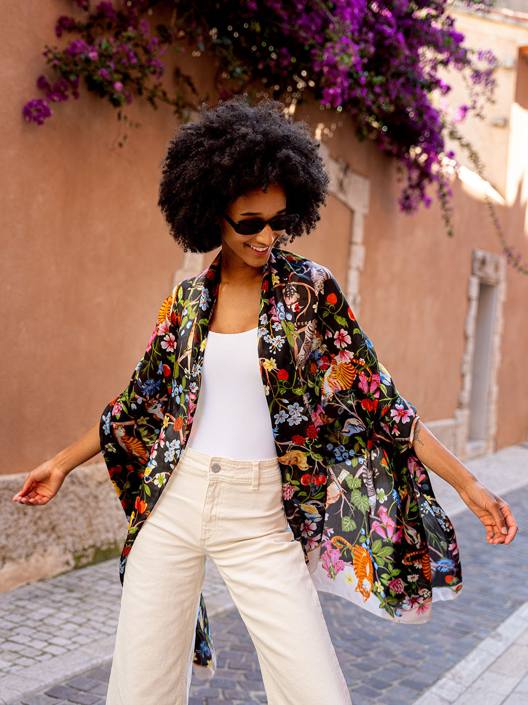 Frau mit Sonnenbrille hat sich die Seiden-Stola 622009 um die Schultern gelegt und läuft an Häusern vorbei.