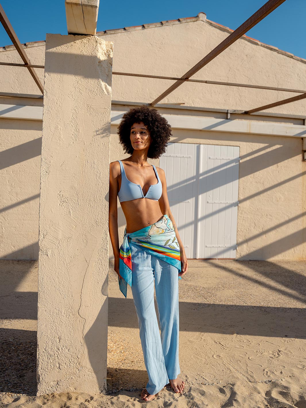 Frau am Strand hat sich das Seidentuch 633037 als Rock umgebunden und steht an eine Säule gelehnt da.