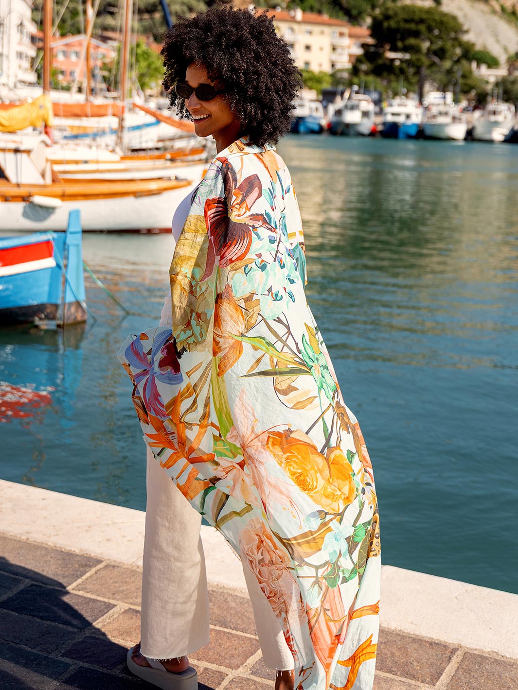 Frau hat sich die FRAAS Seidenstola 609063 um die Schultern gelegt und läuft durch den Hafen, im Hintergrund schaukeln Boote auf dem Wasser.