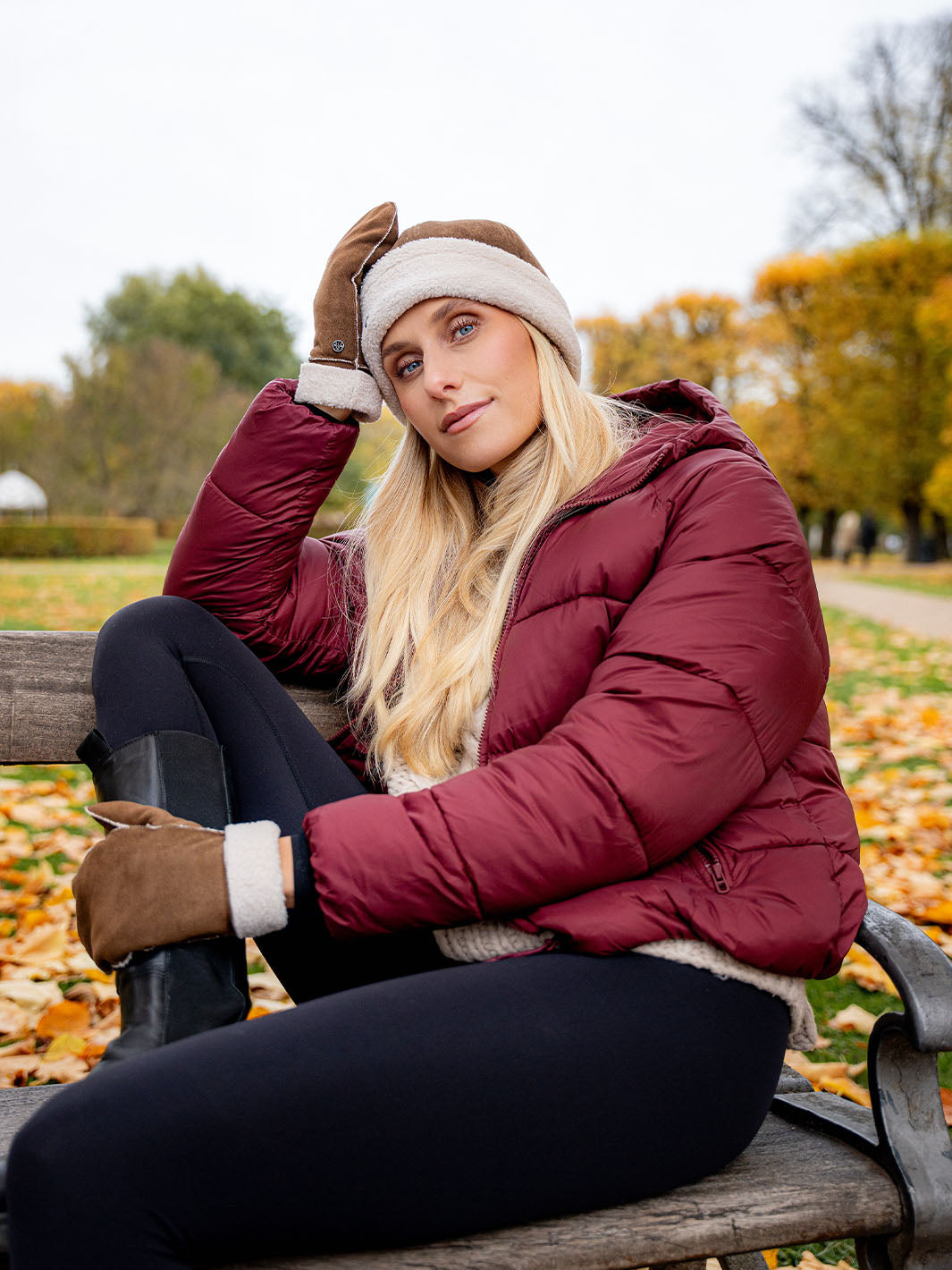Frau in warmer Kleidung sitzt im Park auf einer Bank und hat den Kopf in die Hand gestützt.