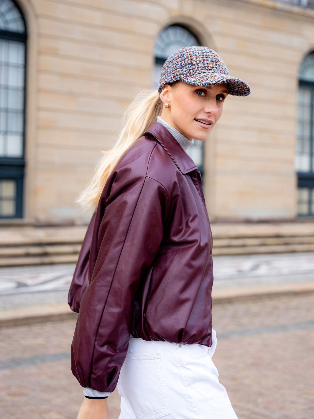 Frau mit Loevenich Basecap aus Tweed mit dezentem Glitzer 679034 geht an einem Haus mit bodentiefen Fenstern vorbei.