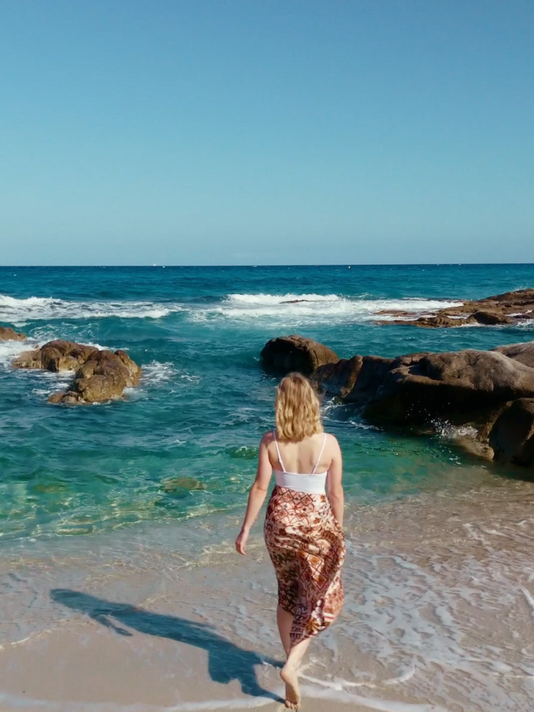 Frau läuft mit dem Rücken zur Kamera zum Meer. Mit den Füßen steht sie schon in den flachen Wellen.