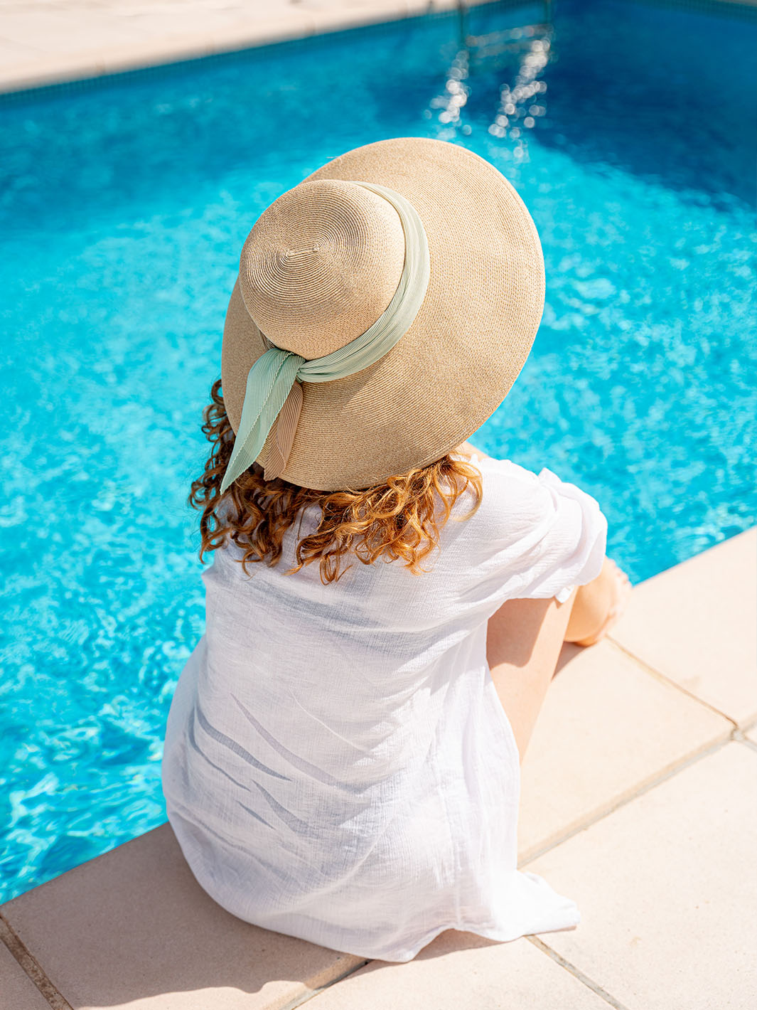 Frau mit Sonnenhut auf dem Kopf sitzt am Pool.