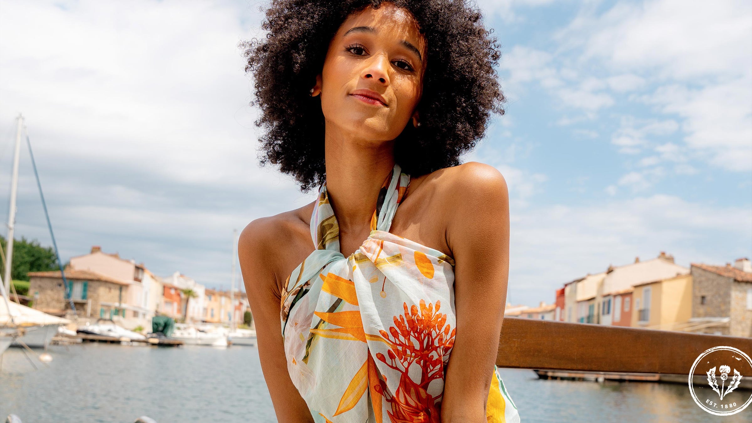Frau trägt die FRAAS Seidenstola 609063 als Top. sie sitzt auf einer Mauer, hinter ihr das Meer, ein Hafen mit Booten und Häuser.