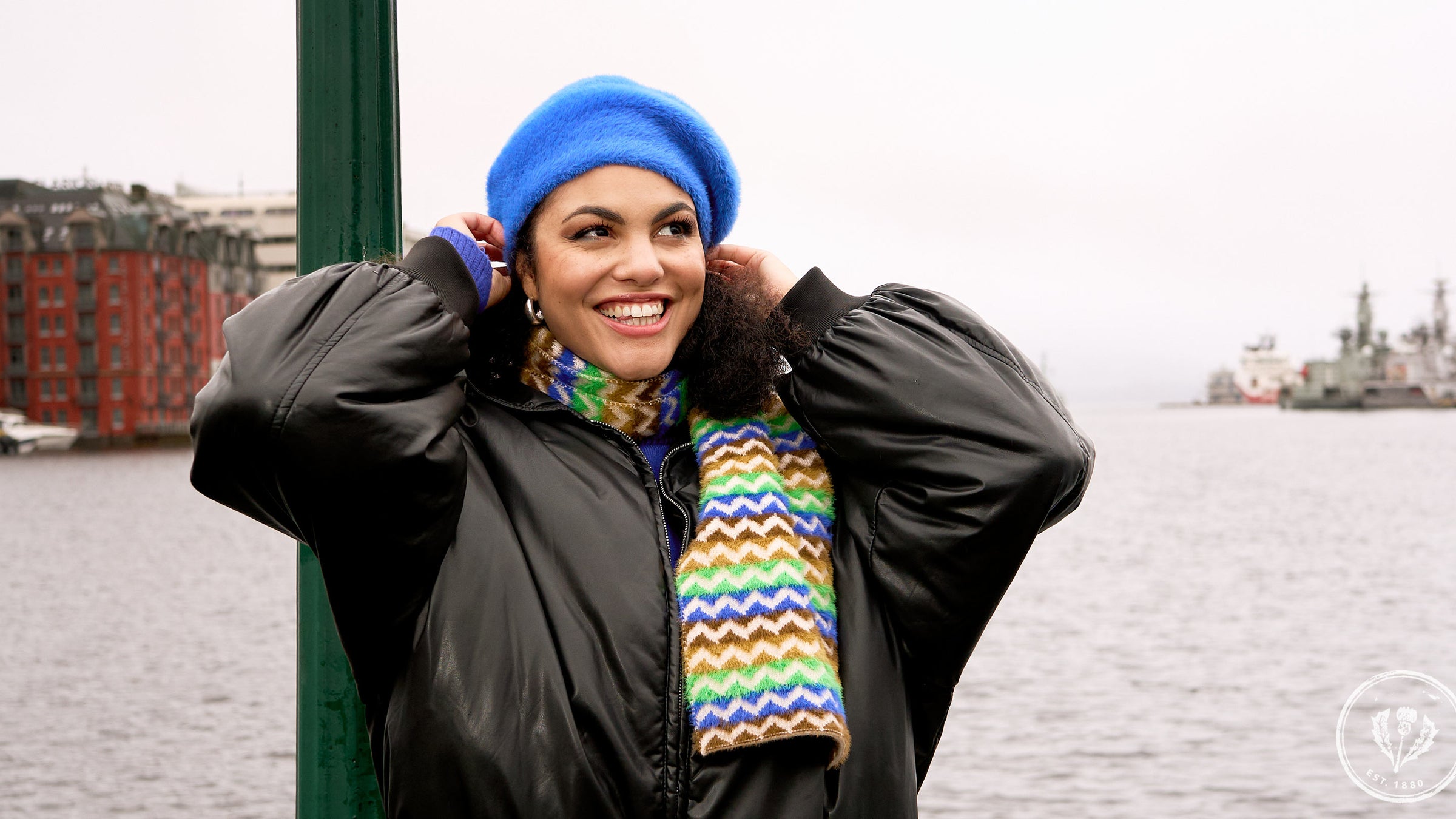 Lachende Frau mit Baskenmütze und dazu passendem Schal steht vor einem Hafen, im Hintergrund Wasser