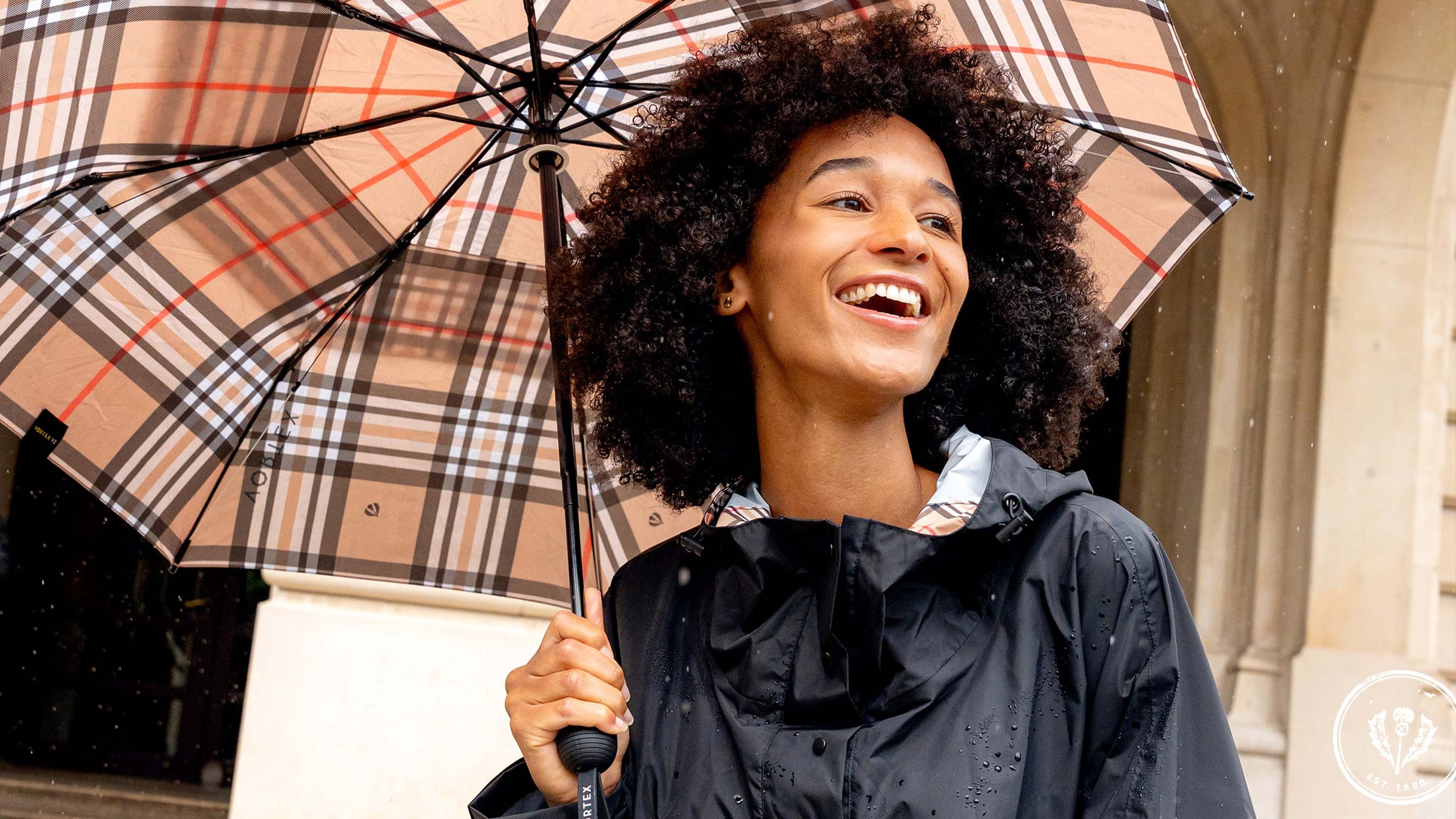 Frau steht lachend unter einem karierten Regenschirm und trägt den FRAAS Plaid Regen-Poncho 602801