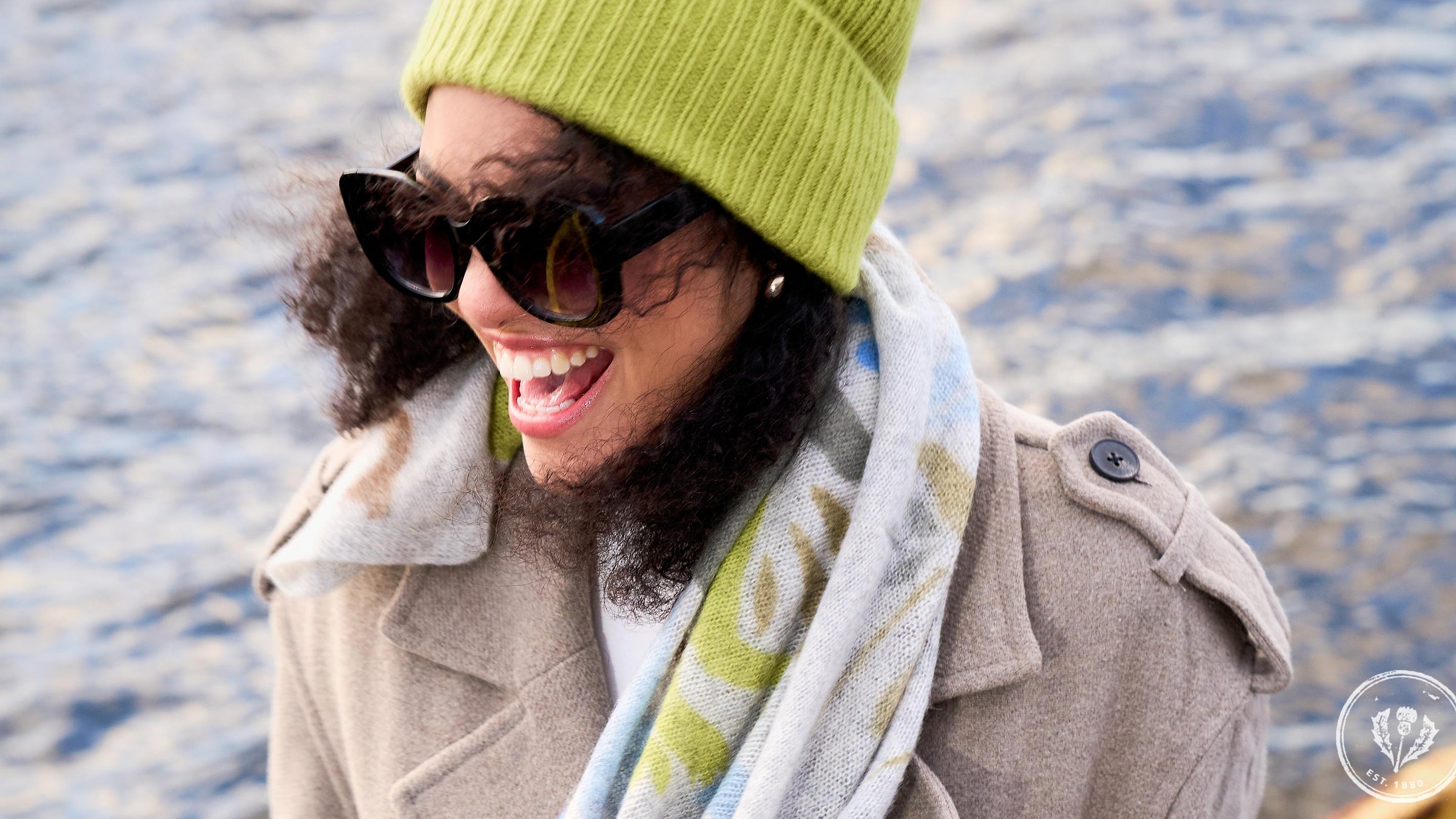 Nahaufnahme lachende Frau mit Sonnenbrille, warmer Wintermütze und Schal vor dem Meer