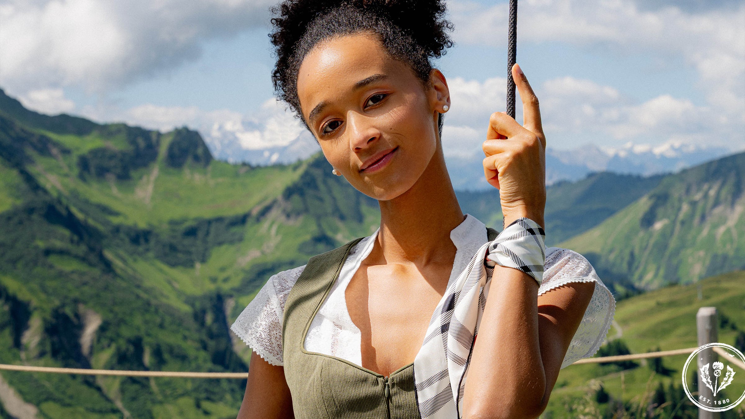 Frau im Dirndl sitzt auf einer Schaukel; im Hintergrund Berge