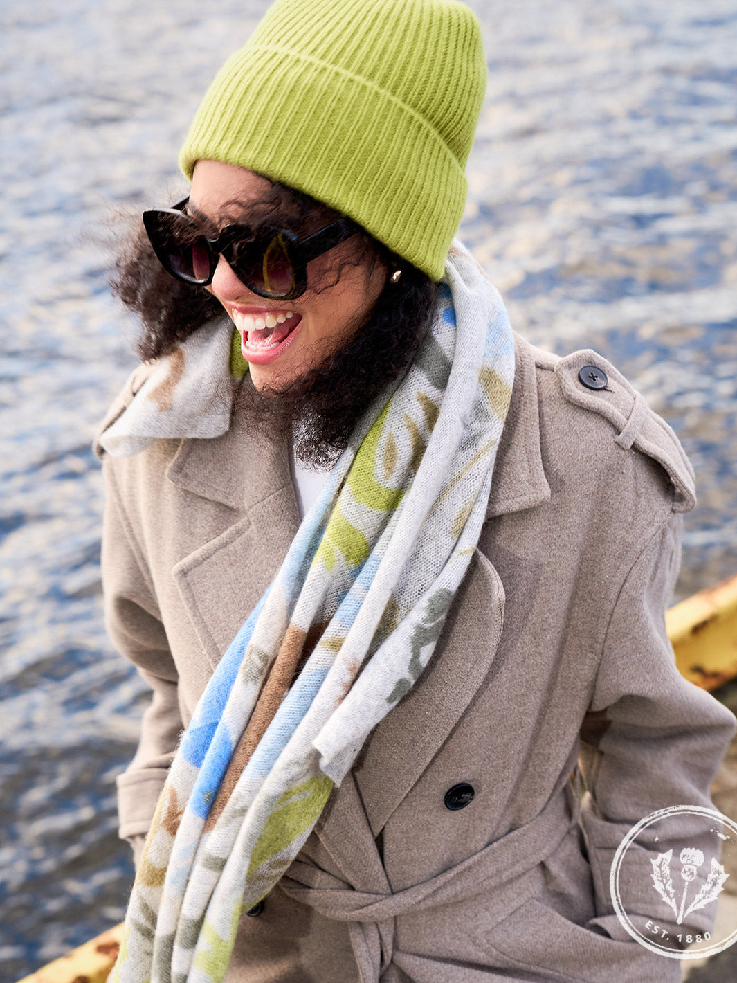 Nahaufnahme lachende Frau mit Sonnenbrille, warmer Wintermütze und Schal vor dem Meer