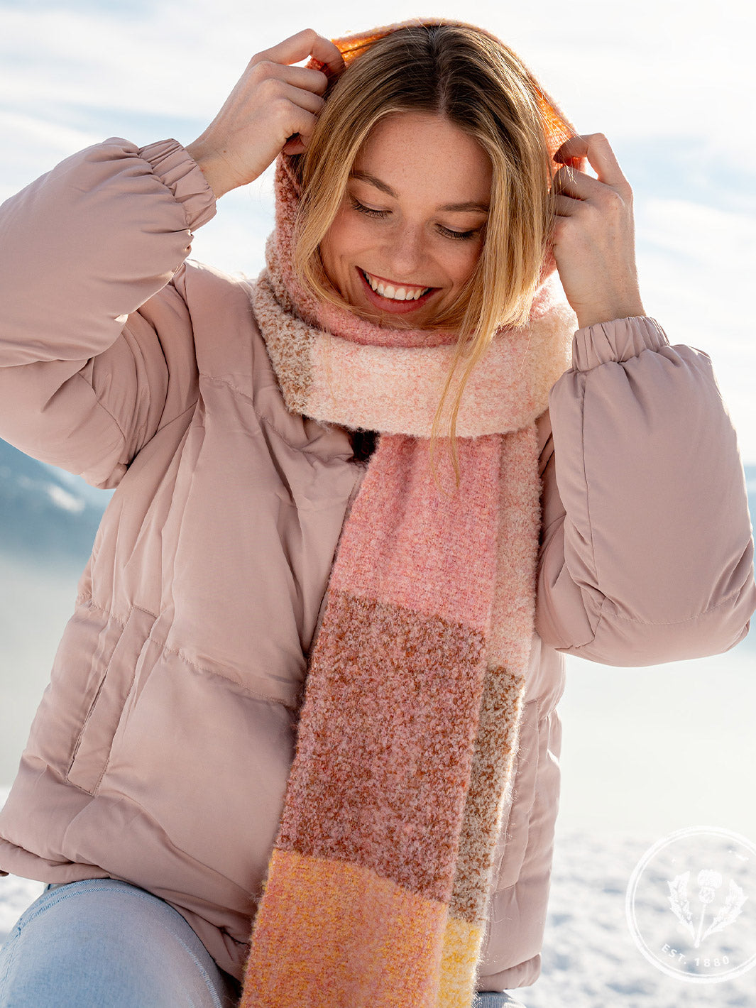 Lächelnde Frau in Winterjacke zieht sich ihren Schal als Mütze über den Kopf