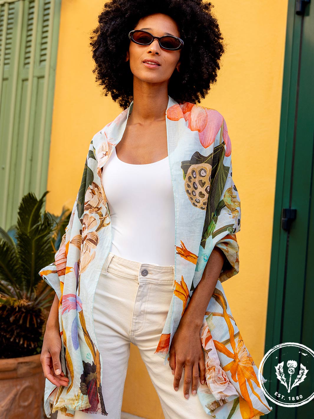 Frau mit Sonnenbrille und Seidenstola 609063 steht vor einer bunten Hauswand und Fensterläden und sieht in die Kamera.