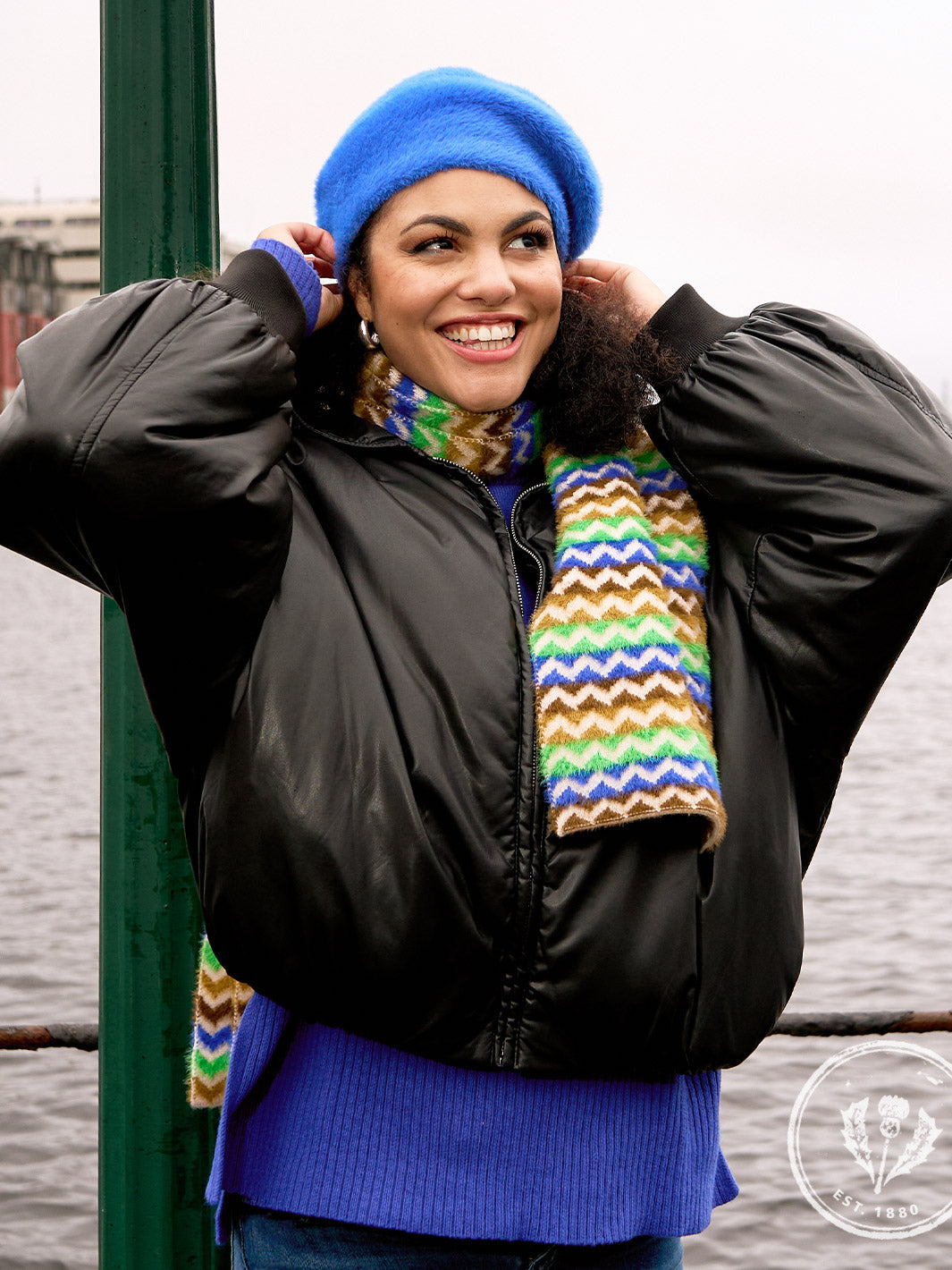 Lachende Frau mit Baskenmütze und dazu passendem Schal steht vor einem Hafen, im Hintergrund Wasser