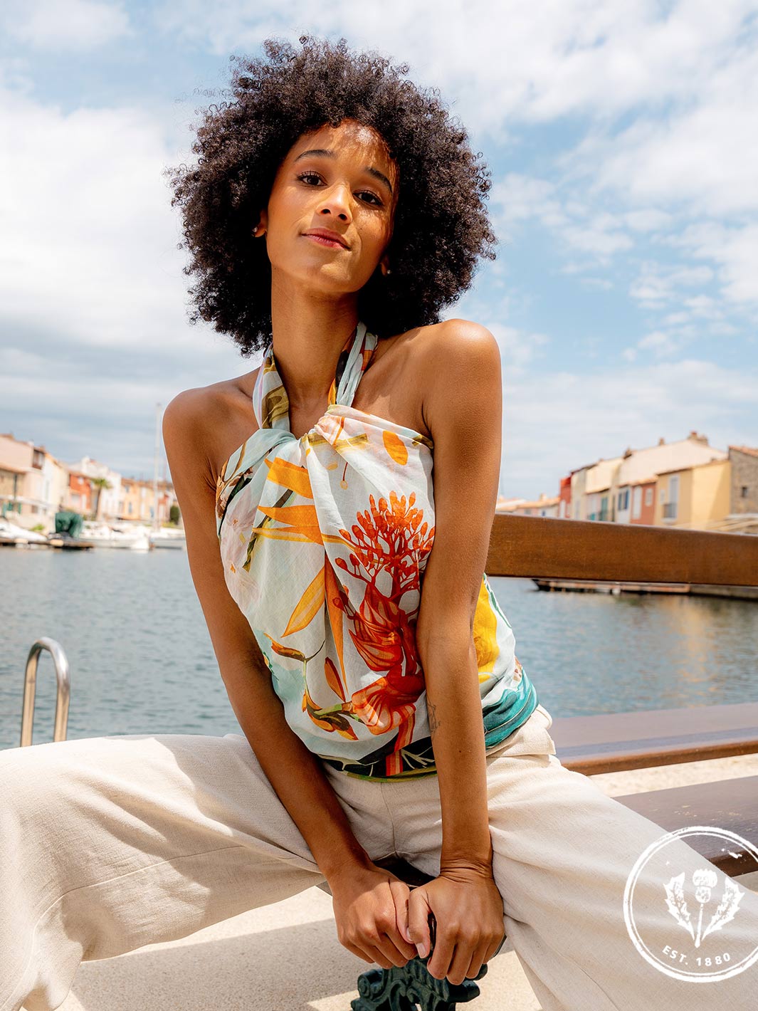 Frau trägt die FRAAS Seidenstola 609063 als Top. sie sitzt auf einer Mauer, hinter ihr das Meer, ein Hafen mit Booten und Häuser.