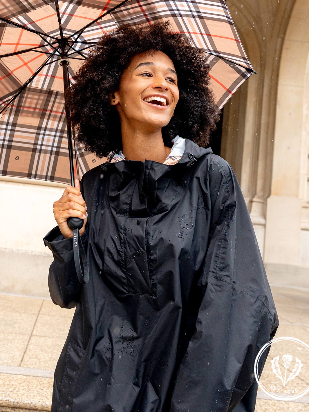 Frau steht lachend unter einem karierten Regenschirm und trägt den FRAAS Plaid Regen-Poncho 602801