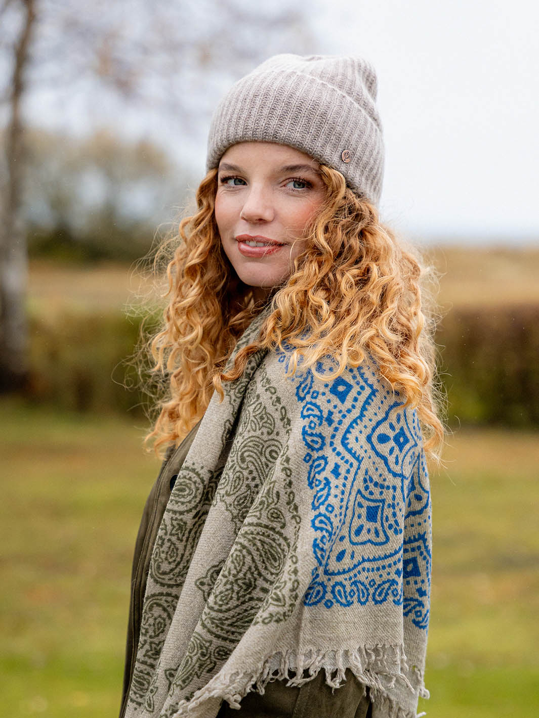 Nahaufnahme einer jungen Frau mit Locken, die mit Strickmütze und FRAAS Schultertuch auf einer herbstlichen Wiese steht.